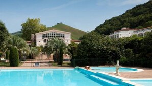 A large swimming pool with a fountain, surrounded by lush greenery and a hill in the background. - STUDIO GETTE HASPARREN in the Basque Country near the Cambo thermal baths (Hasparren)