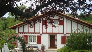 A two-story house with a white exterior and red trim, featuring a front porch with a bench and potted plants. - STUDIO GETTE HASPARREN in the Basque Country near the Cambo thermal baths (Hasparren)