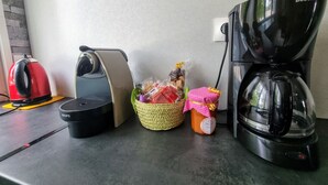 A kitchen counter with a red kettle, a coffee maker, a Krups electric kettle, a woven basket with food items, and a polka-dotted container.