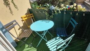 A small outdoor patio with a round table and two chairs, surrounded by a green fence with plants and flowers.
