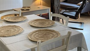 Un coin repas avec une table dressée pour quatre personnes, une structure en poutres apparentes, un canapé et une fenêtre donnant sur un paysage verdoyant.