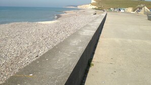 Une plage de galets avec une promenade en béton, un ciel dégagé et des falaises lointaines.