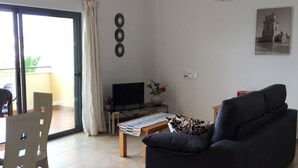 Ein Wohnzimmer mit Sofa, Fernseher, Tisch und Stuhl. Eine Schiebetür führt zu einem Balkon.