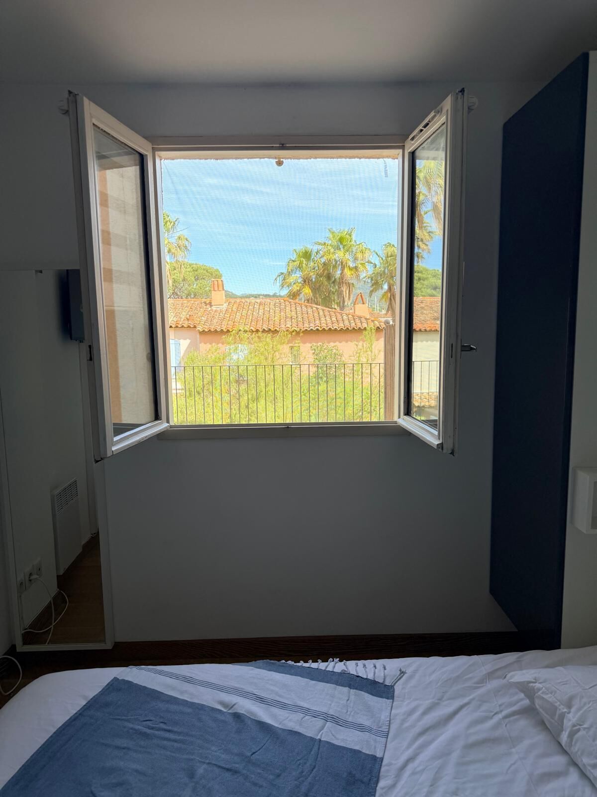 Une chambre avec une grande fenêtre, un lit recouvert d’une couverture à rayures bleues et blanches, et une vue sur un bâtiment doté d’un toit en tuiles.