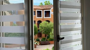 An open window with horizontal blinds, a view of a courtyard with a bench, and a building with a tiled roof. - 2 Bedroom apartment with swimming pool and underground parking (Grimaud)
