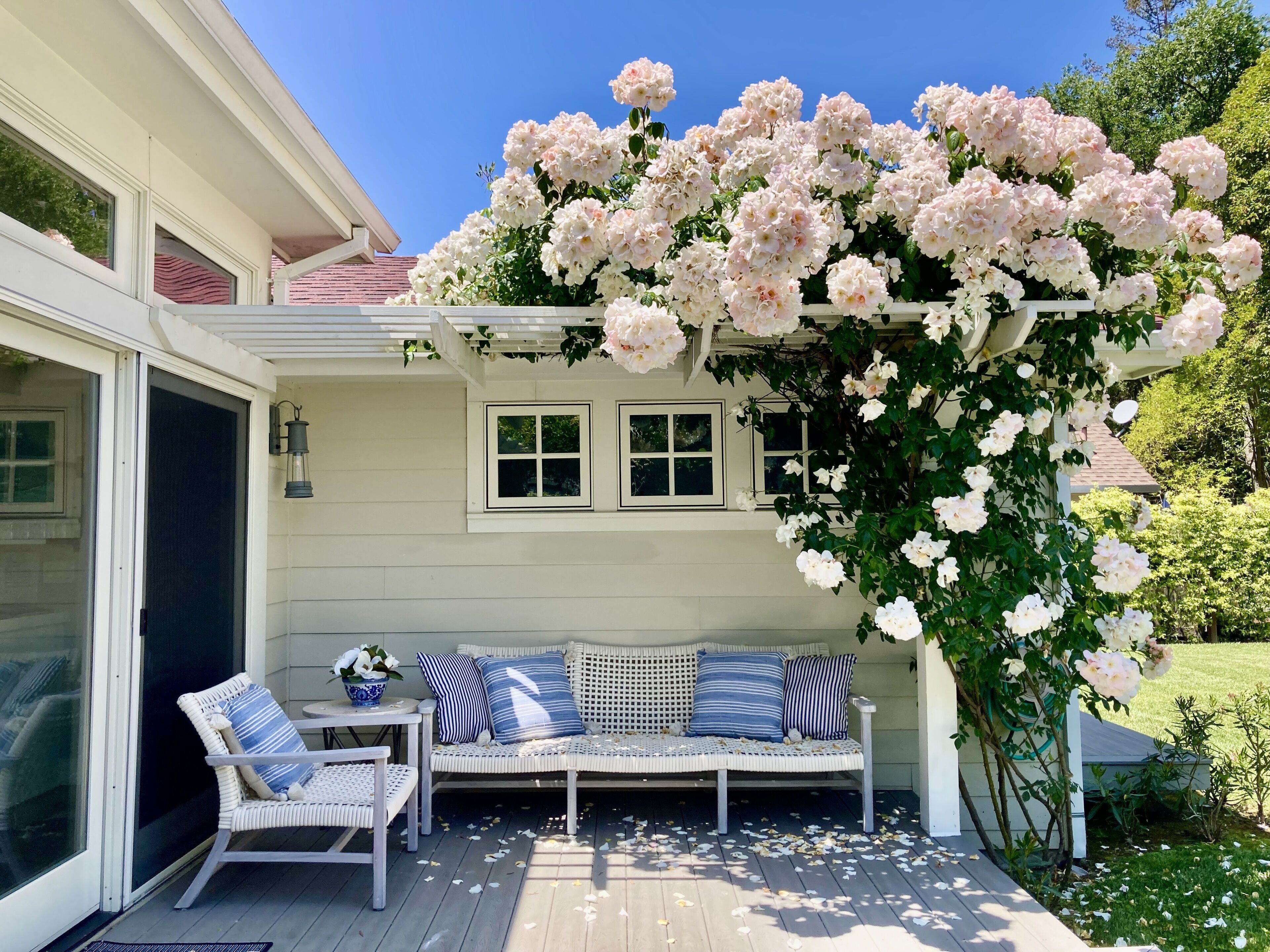 A covered patio with a white bench and chairs, surrounded by blooming pink flowers.