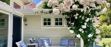 A covered patio with a white bench and chairs, surrounded by blooming pink flowers.
