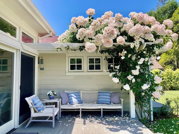 A covered outdoor seating area with a white bench and chairs, surrounded by a lush garden with flowering plants.