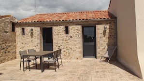 Un edificio de piedra con techo de tejas, una mesa de comedor con sillas, y un pequeño espacio exterior con una silla y una mesa.