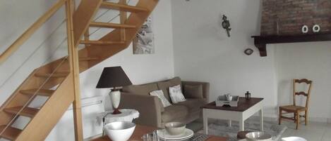 Un comedor con una mesa de madera dispuesta para cuatro personas, una escalera, un sofá y una pared de ladrillo.