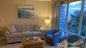 A living room with a sofa, two armchairs, a coffee table, and a lamp. There is a painting on the wall and a window with a view of greenery and a building. - Villamare 3328, New pools, Ocean views, WiFi, Beachfront, beautifully remodeled. (Hilton Head Island)