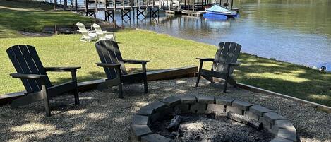 Un foc de campament envoltat de cadiretes Adirondack, un moll amb vaixells i un mirador al costat d'un llac.