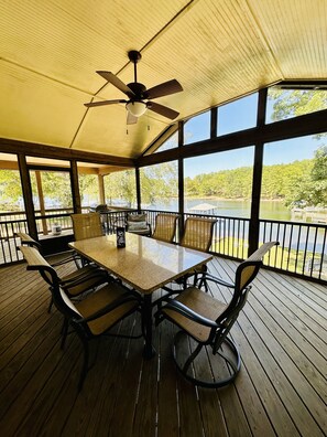 A covered outdoor dining area with a table and chairs, large windows, and a view of a lake.