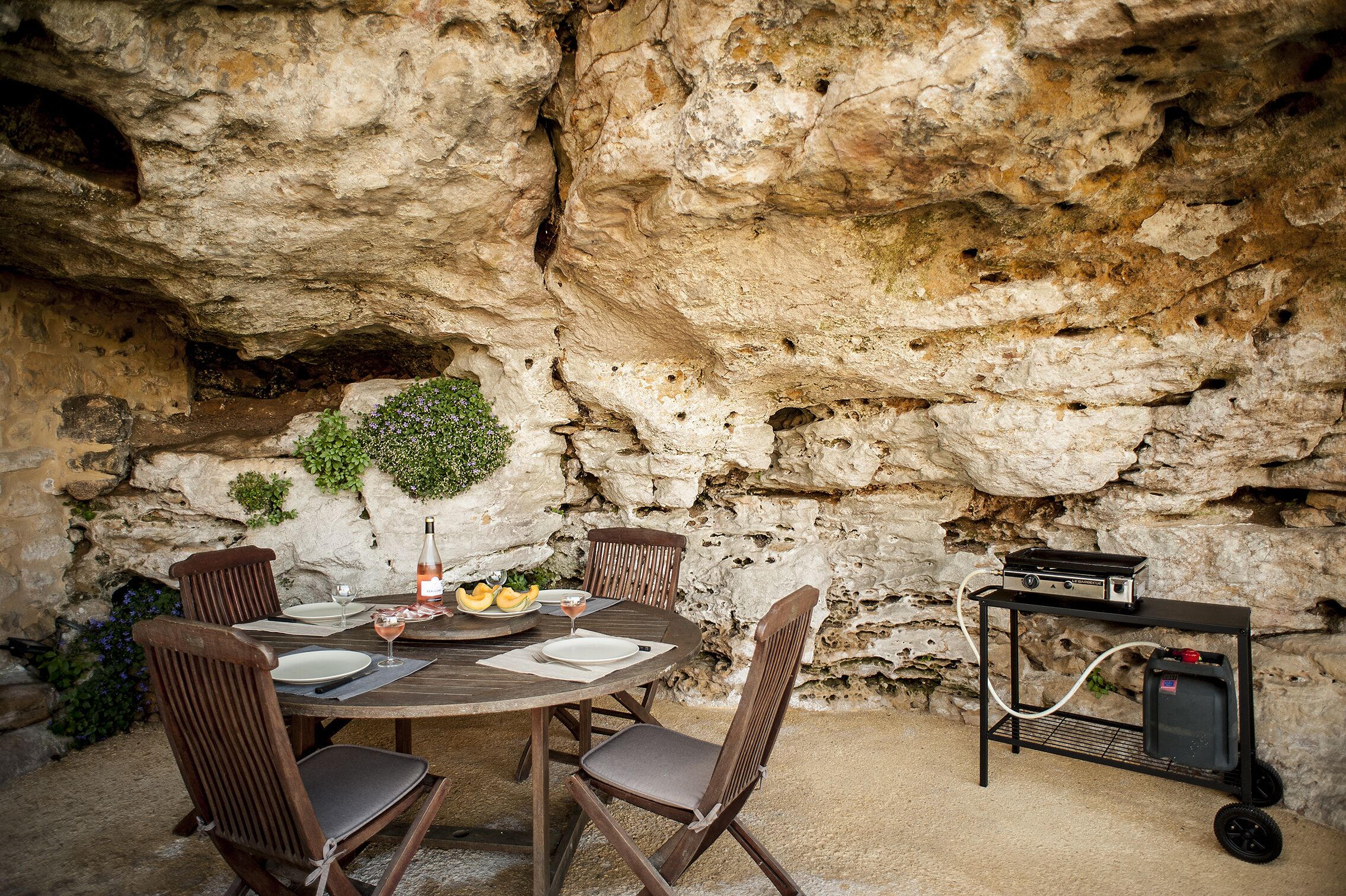 A rustic outdoor dining area with a stone wall backdrop, a round wooden table set for four, and a portable grill.