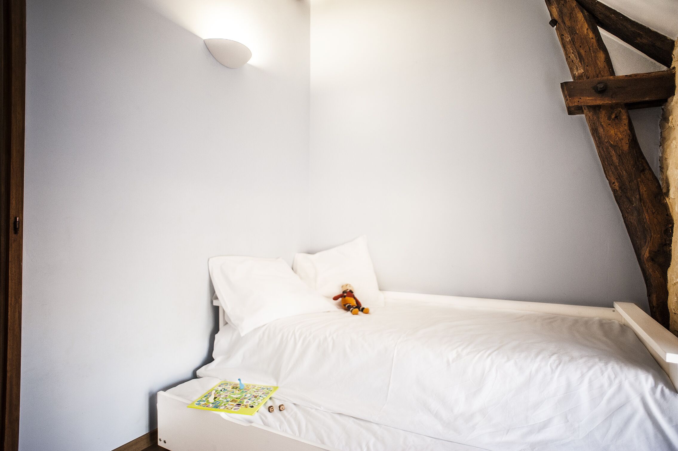 Quarto com cama branca, cabeceira de madeira e luminária fixa na parede.