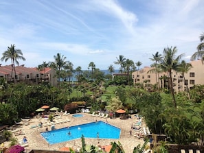 A resort with a central pool surrounded by lounge chairs and umbrellas, palm trees, and buildings with red roofs.