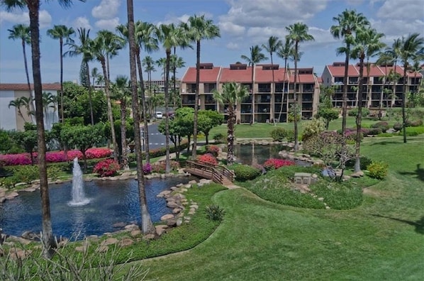 A hotel with a landscaped garden, palm trees, a pond with a fountain, and a parking area.