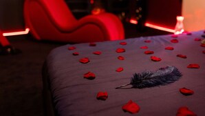 Ein Bett mit Rosenblättern und einer Feder, ein roter Stuhl und eine brennende Kerze in einem schwach beleuchteten Raum.