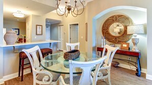 A dining area with a glass table, white chairs, a chandelier, and a decorative mirror. - Luxury Condominium Next Door to the Ritz Carlton  (Fernandina Beach)