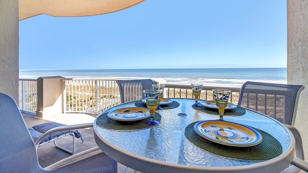 A round table with a glass top, surrounded by chairs, set for dining with plates and glasses, overlooking a beach with the ocean in the background. - Luxury Condominium Next Door to the Ritz Carlton  (Fernandina Beach)