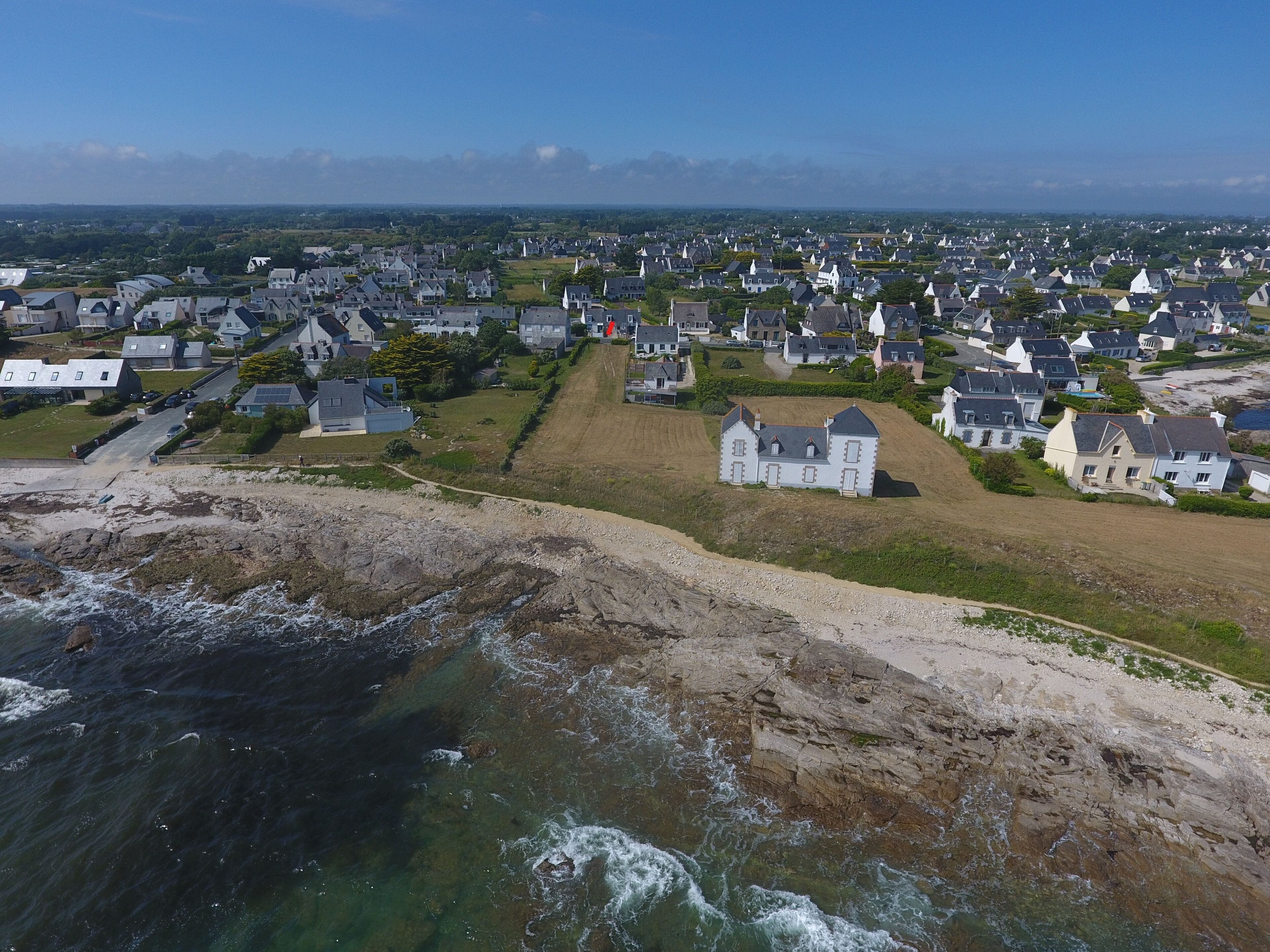 Vue aérienne d’une zone résidentielle côtière, avec des maisons, un grand bâtiment blanc et une côte rocheuse.