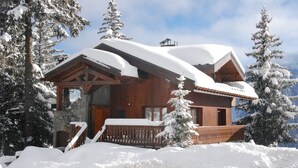 Une cabane en bois, posée sur des fondations en pierre, entourée d’arbres couverts de neige et dotée d’une terrasse en bois.