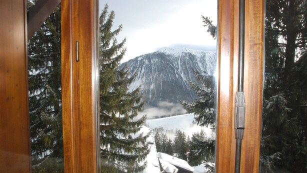 Une vue à travers une fenêtre à cadre en bois, donnant sur un paysage montagneux avec des arbres couverts de neige et un lac.