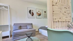 A modern bedroom with a grey sofa, a round coffee table, and a bed with patterned bedding. There are three framed artworks on the wall and a wooden panel on the right.
