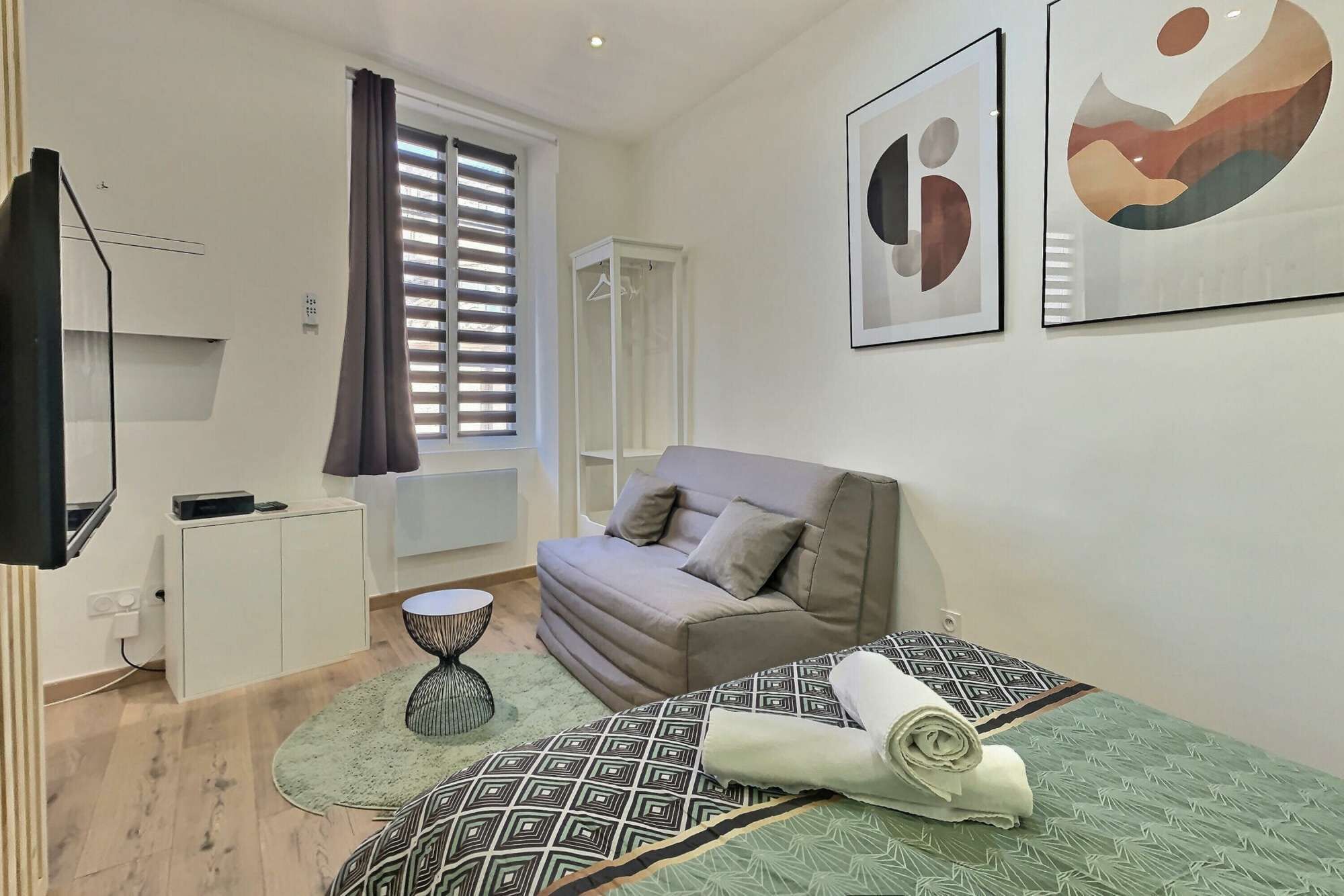 A modern bedroom with a sofa, a small round table, a bed with a patterned bedspread, and abstract wall art.