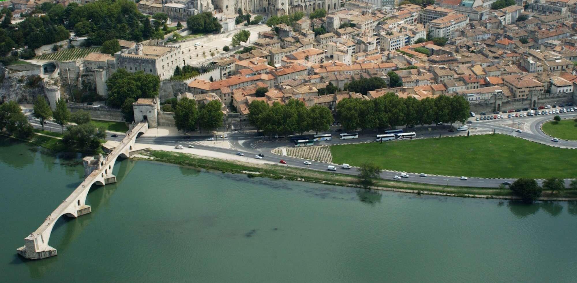 An aerial view of Pont d'Avignon with a medieval bridge over a river, surrounded by historic buildings and a pedestrian path.