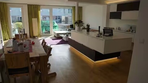 Ett modernt kök med en köksö i mitten, en matplats med ett träbord och stora fönster med gardiner.