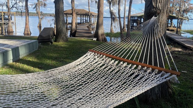 A hammock tied to a tree by a wooden dock extending into a lake, surrounded by trees and a gazebo.