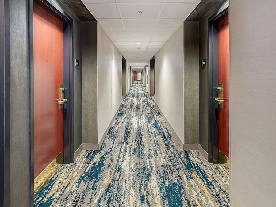 A hotel corridor with carpeted flooring, doors on both sides, and a ceiling with recessed lighting.