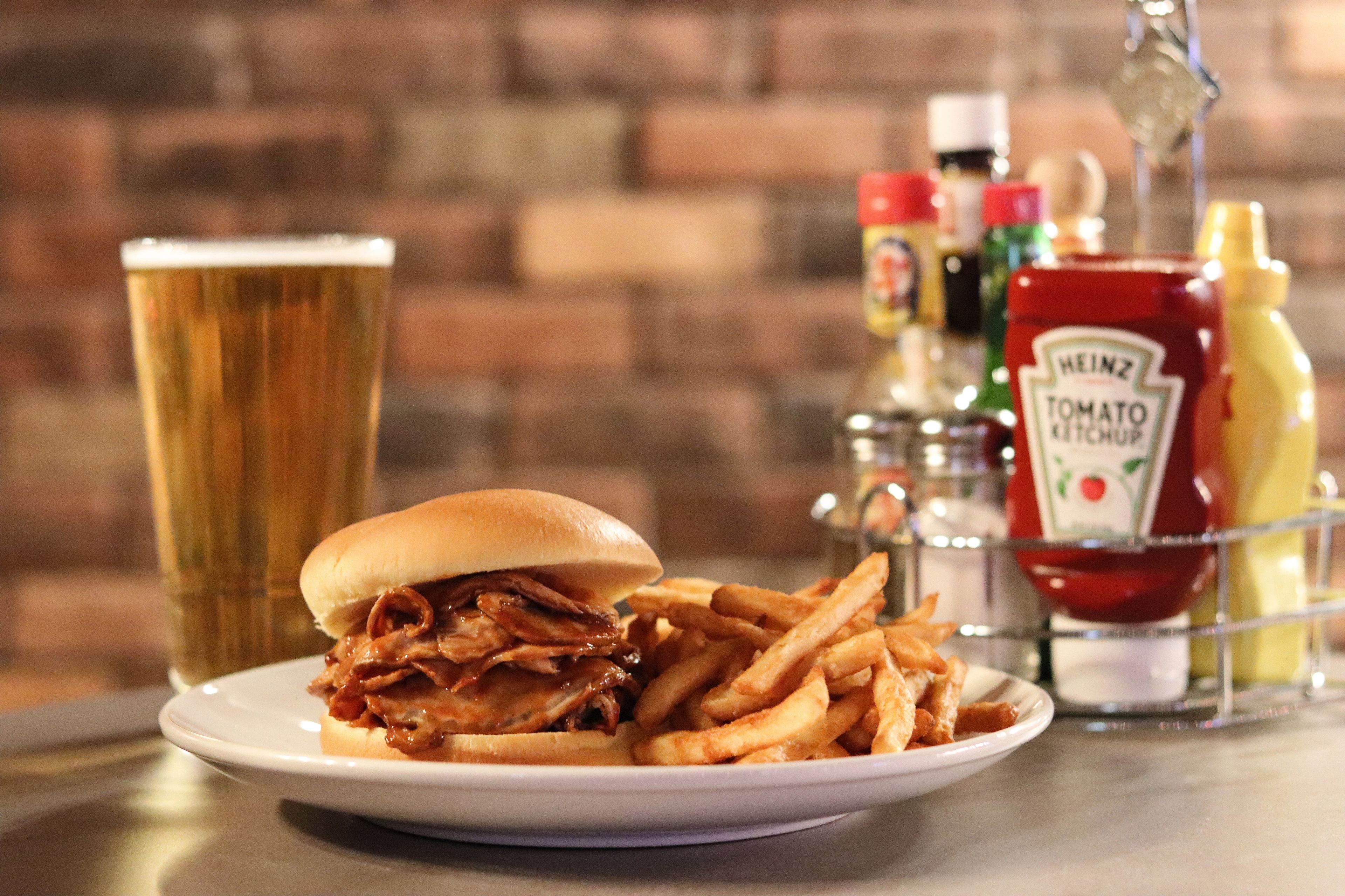 Een bord met een pulled pork sandwich, frieten en een glas bier, met sauzen op tafel.