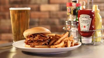 Een bord met een pulled pork sandwich, frieten en een glas bier, met sauzen op tafel.