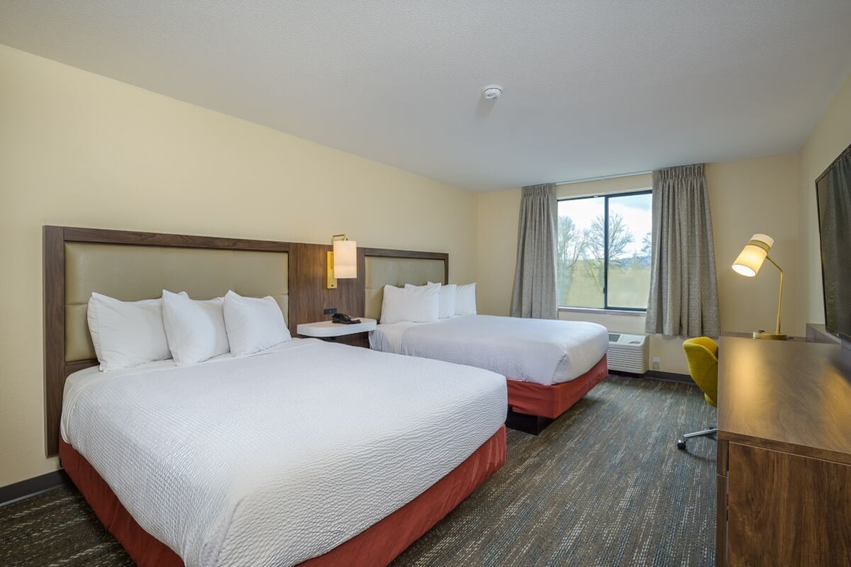 A hotel room with two beds, a desk, a lamp, and a window with curtains.