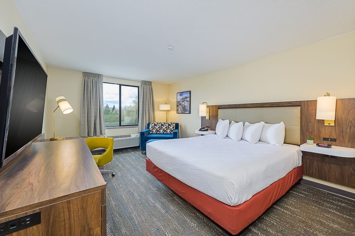 A hotel room with a large bed, a desk with a flat-screen TV, a chair, a window with curtains, and a wall-mounted lamp.