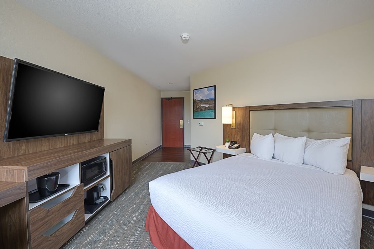 A hotel room with a large bed, a flat-screen TV mounted on a wooden cabinet, a small table with a lamp, and a painting on the wall.