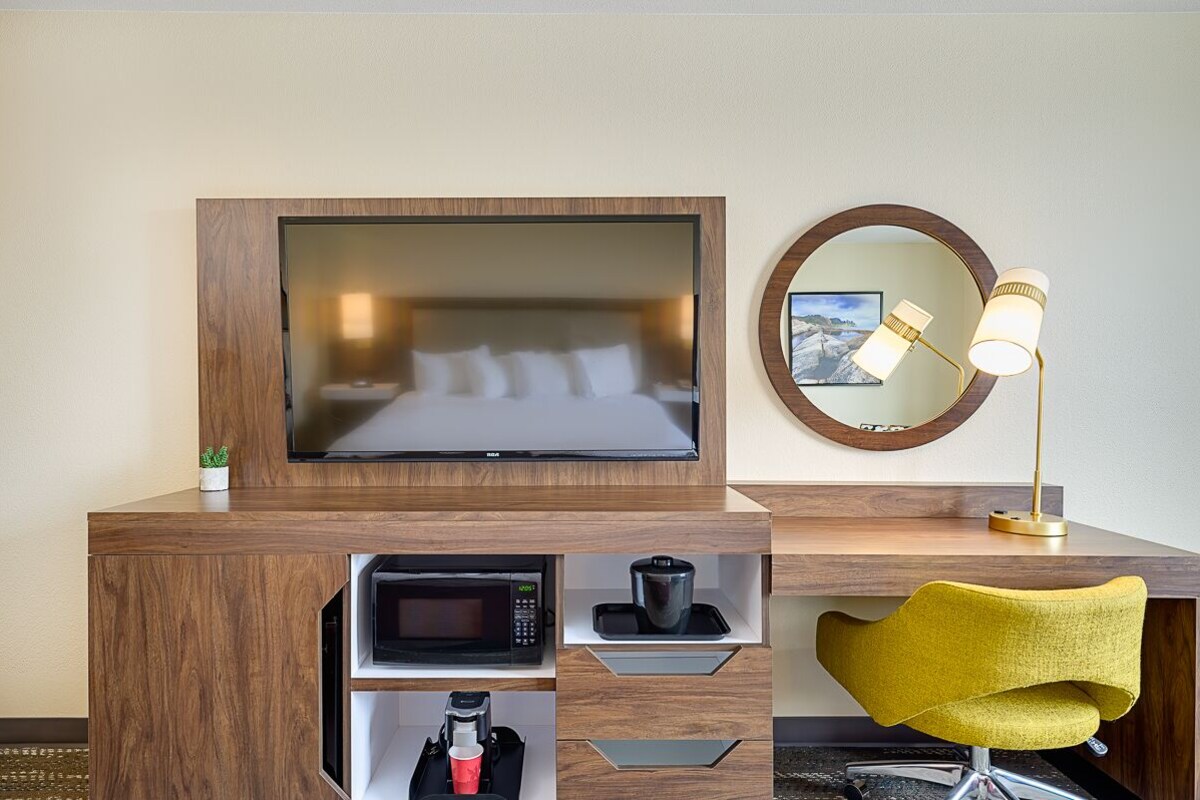 A hotel room with a wooden desk, a television, a coffee maker, a microwave, a mirror, and a yellow chair.