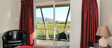 A hotel room with a bed, a chair, a nightstand with a lamp, and a view of a balcony with a railing.