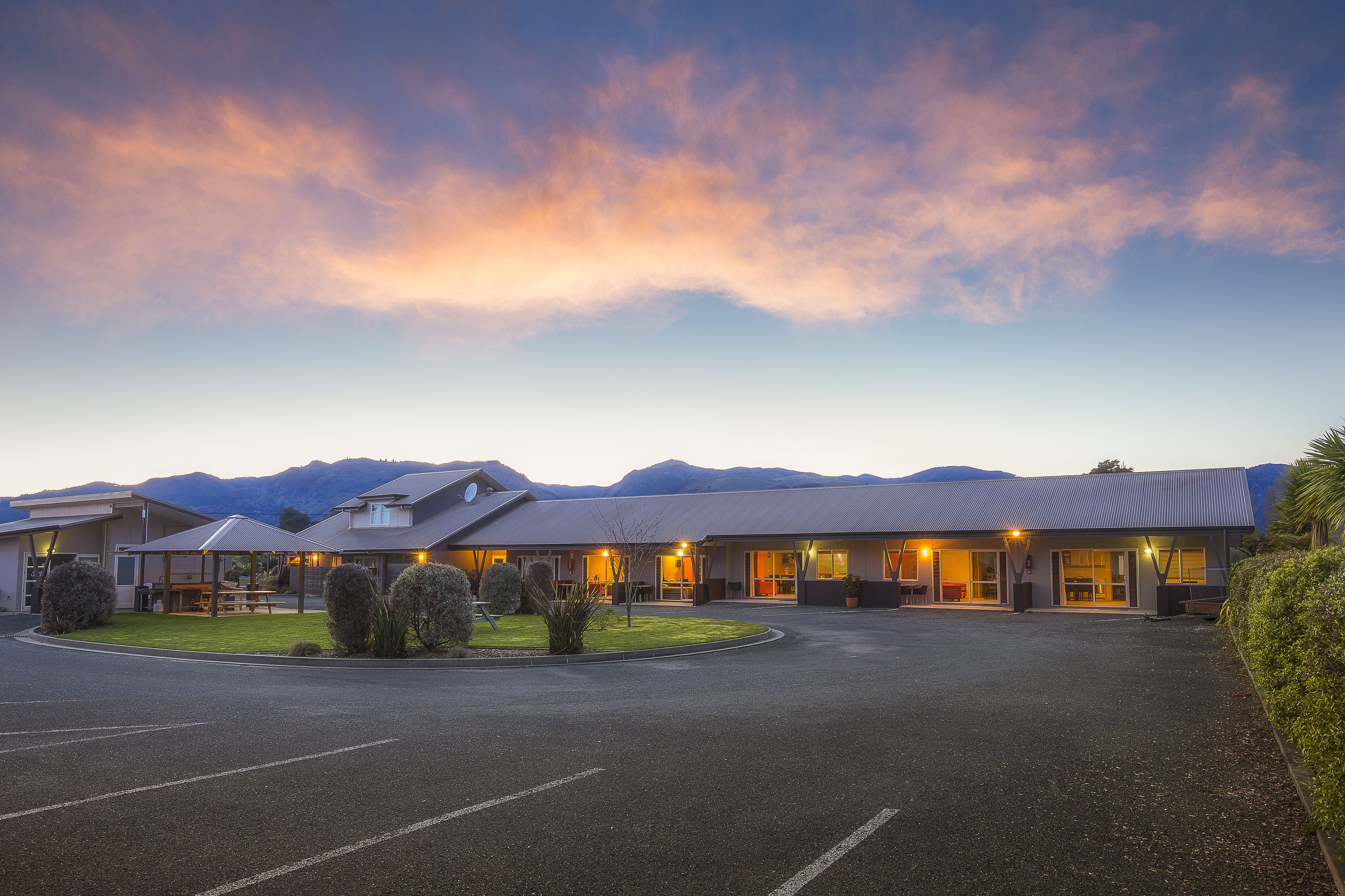 Un hotel con estacionamiento, un área exterior techada y montañas al fondo.