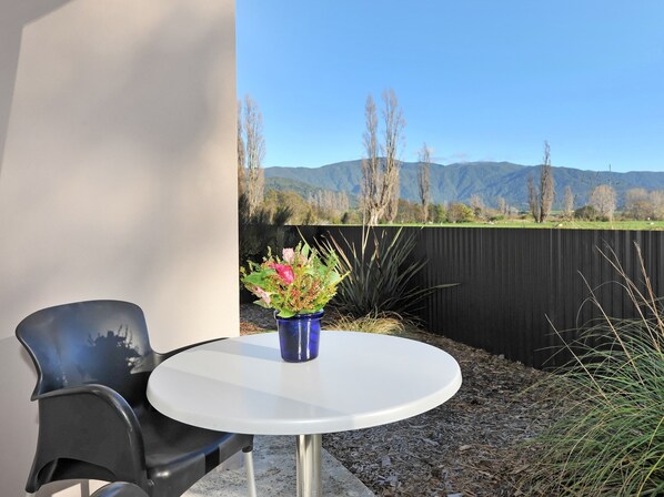Una mesa redonda con un florero azul lleno de flores, una silla negra y una vista de montañas y una cerca al fondo.