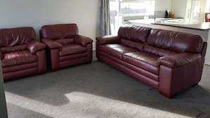 A living room with two brown leather sofas, a kitchen area with white cabinets, and a large window with curtains. - Micro villa (Christchurch)