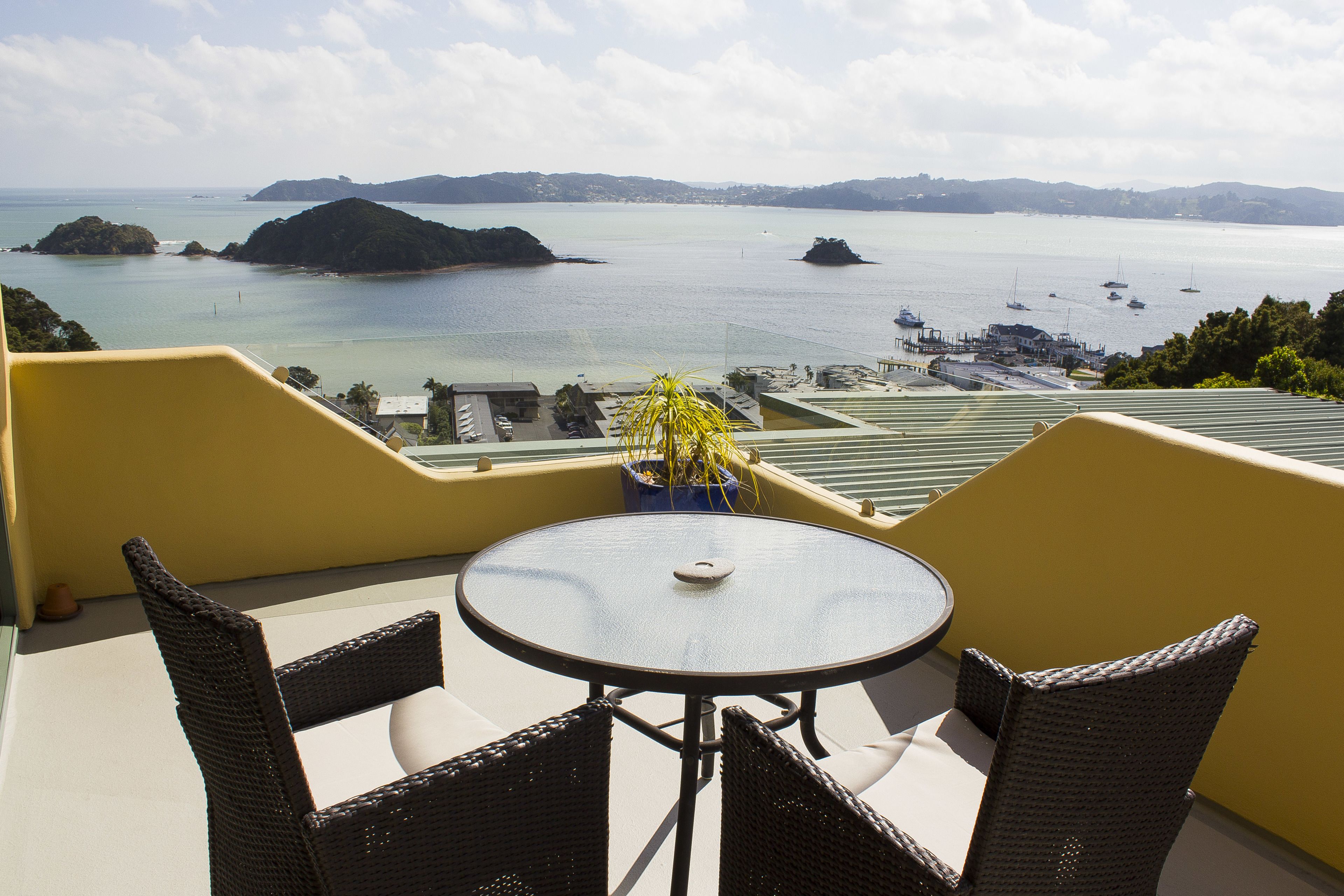 A balcony with wicker chairs and a table, overlooking a bay with boats and distant islands.