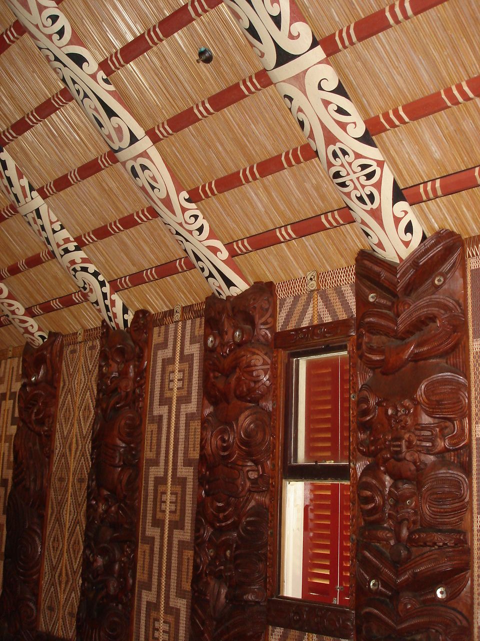 Una habitación con intrincados grabados de madera en el techo y las paredes, techo con motivos decorativos y una ventana con contraventanas rojas.