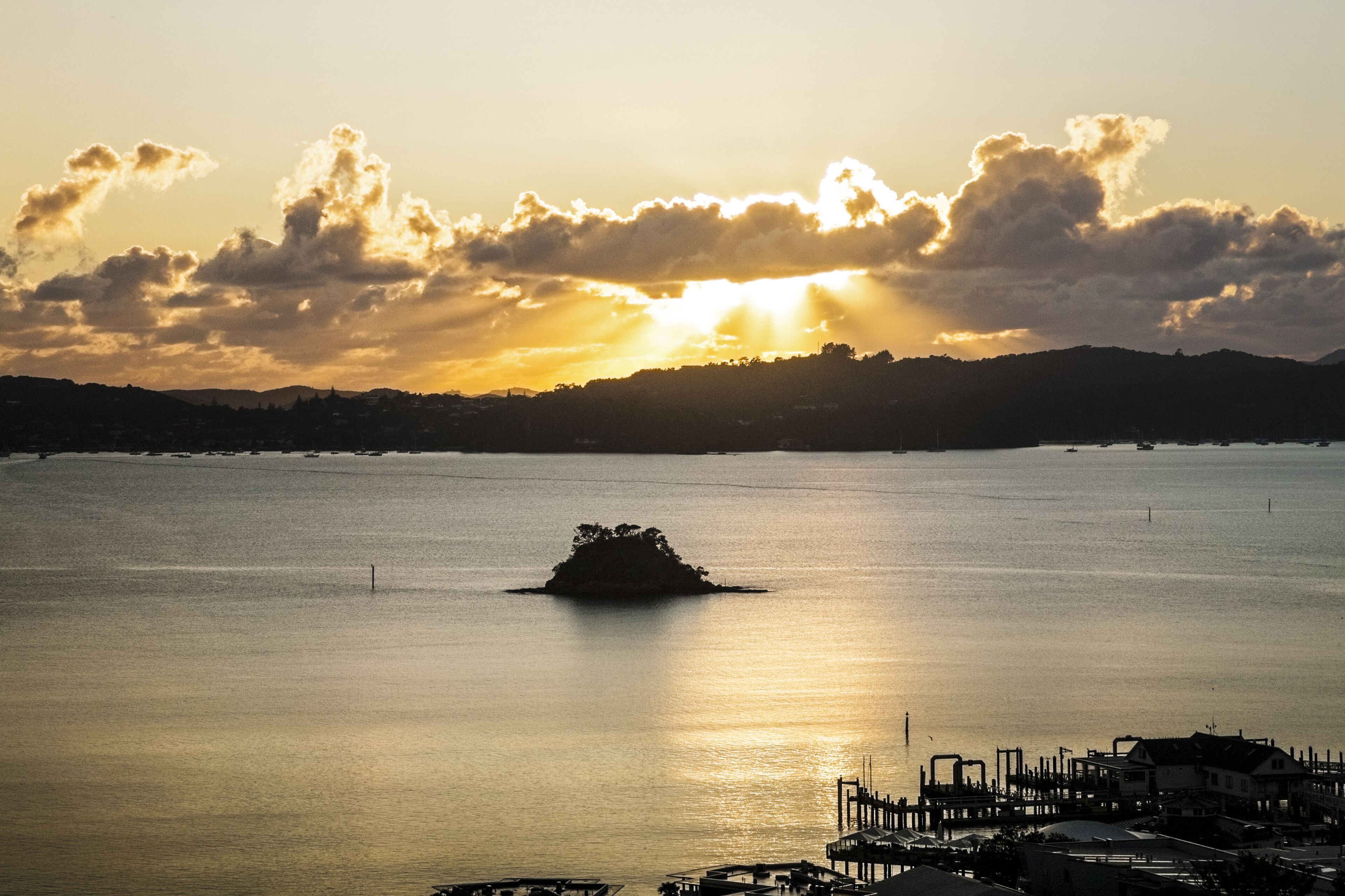 A calm body of water with a small island in the center, a dock area with boats, and a distant shoreline with buildings.