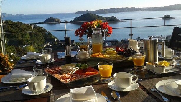 Una mesa de desayuno con vistas al mar y colinas, con platos de fruta, bollería y una jarra de zumo de naranja.