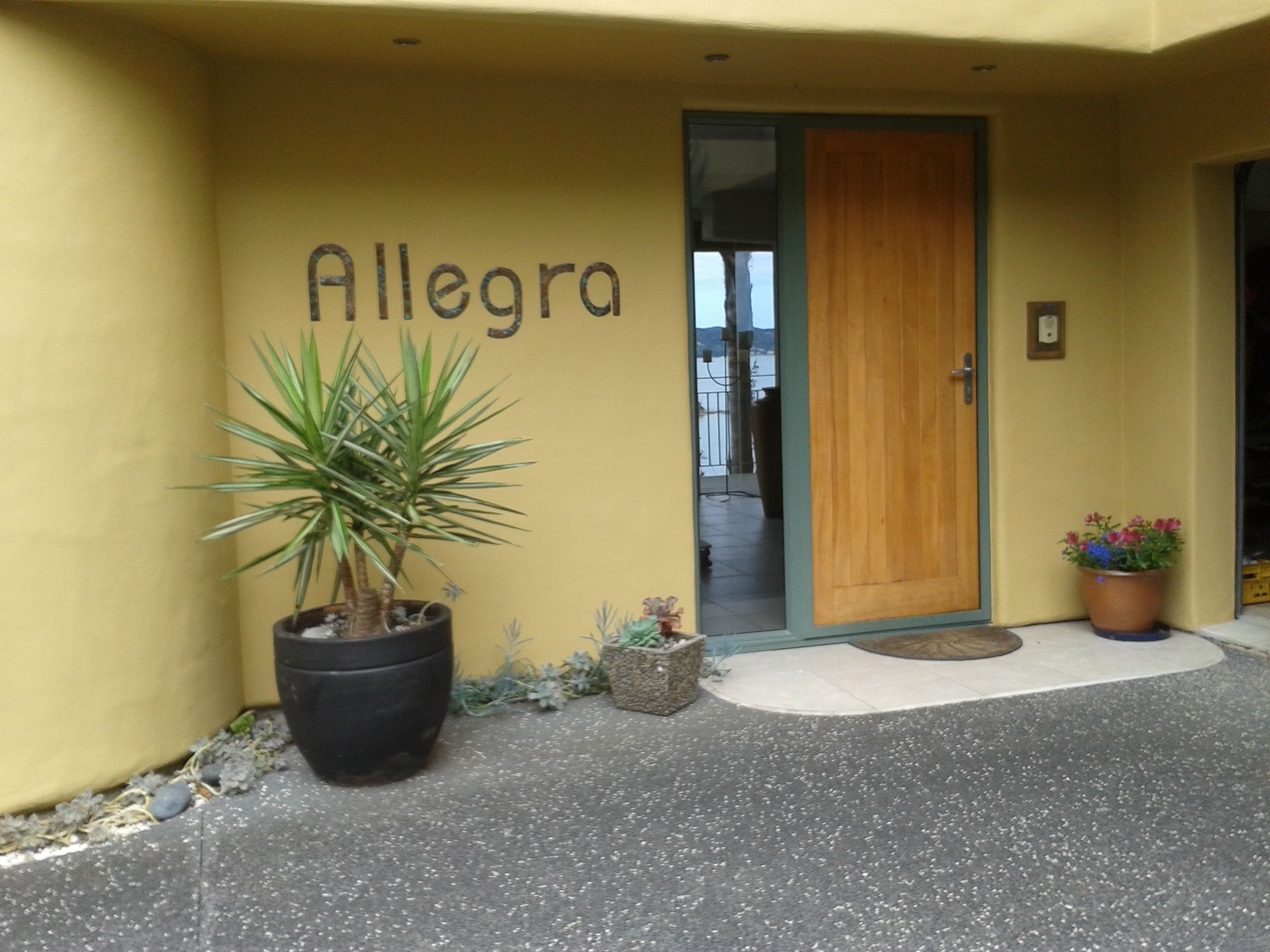 A building entrance with a door labeled "Allegra," a potted plant, and a small garden area.