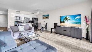 A modern living room with a grey sofa, a glass coffee table, and a flat-screen TV. There is a kitchen area with white cabinets and a dining table with chairs.