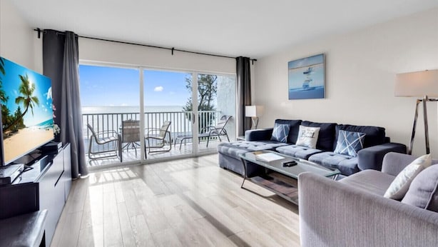 A modern living room with a balcony overlooking the sea, a flat-screen TV, a sofa, and a coffee table.